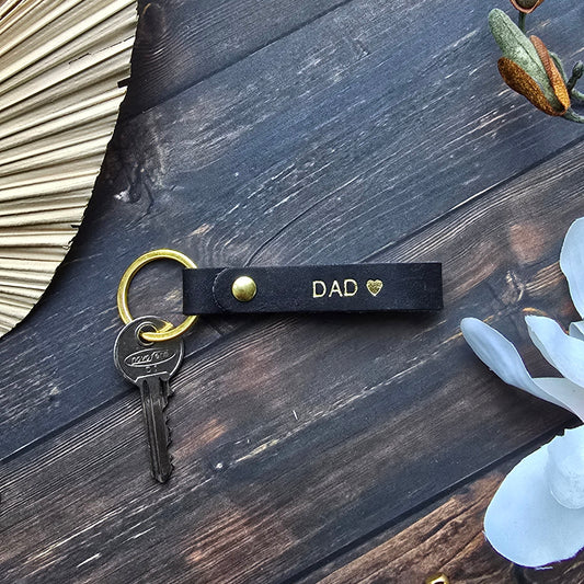 A black leather keychain with gold hardware and the text 'DAD' printed on it, holding a key, styled on a dark wooden background with decorative props.