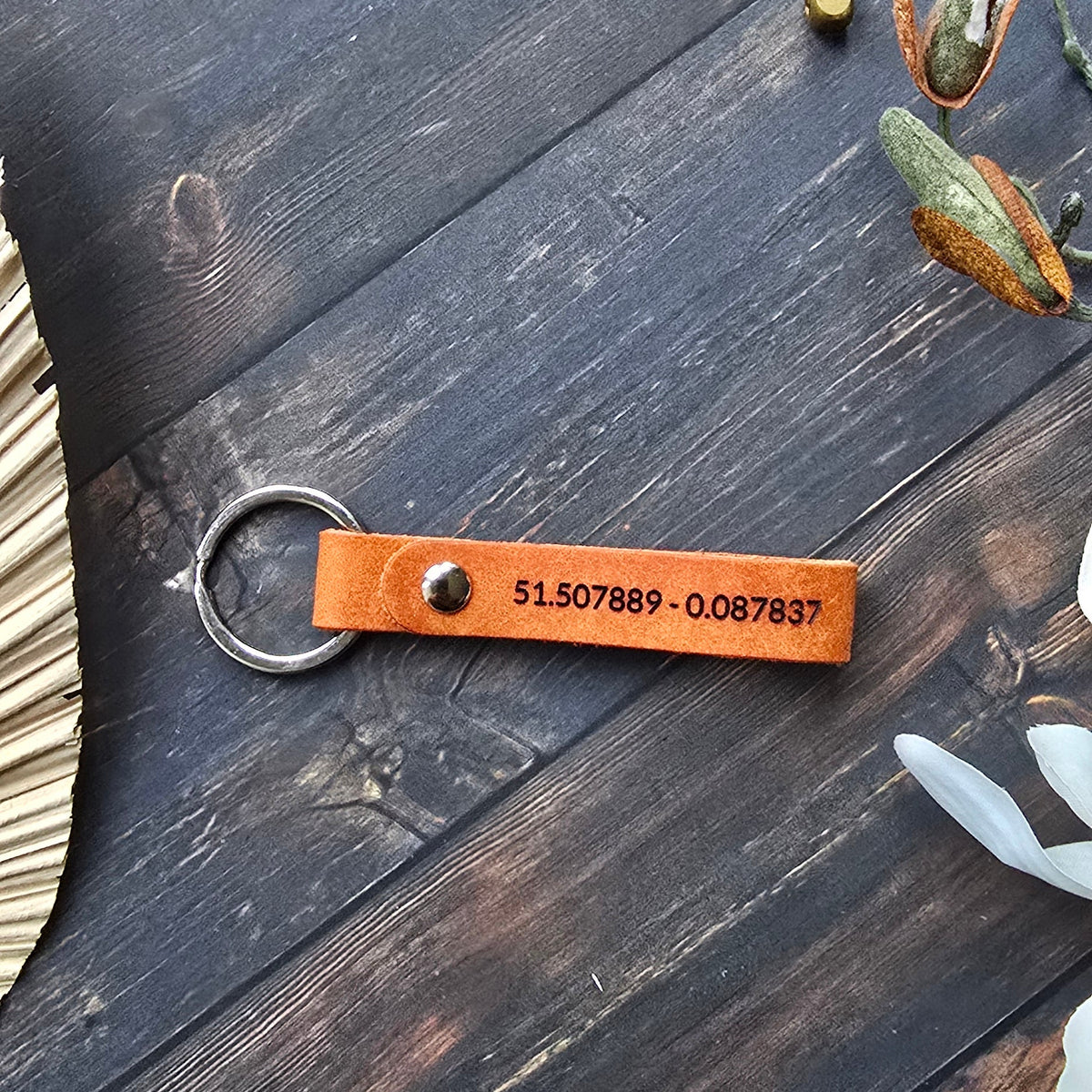 A personalized brown leather keychain with a metal split ring, featuring a custom-engraved phone number, displayed on a dark rustic wooden background with decorative dried leaves and small gold bells.