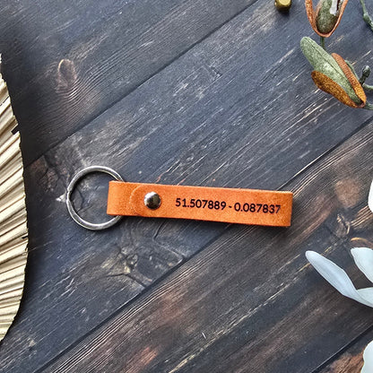 A personalized brown leather keychain with a metal split ring, featuring a custom-engraved phone number, displayed on a dark rustic wooden background with decorative dried leaves and small gold bells.
