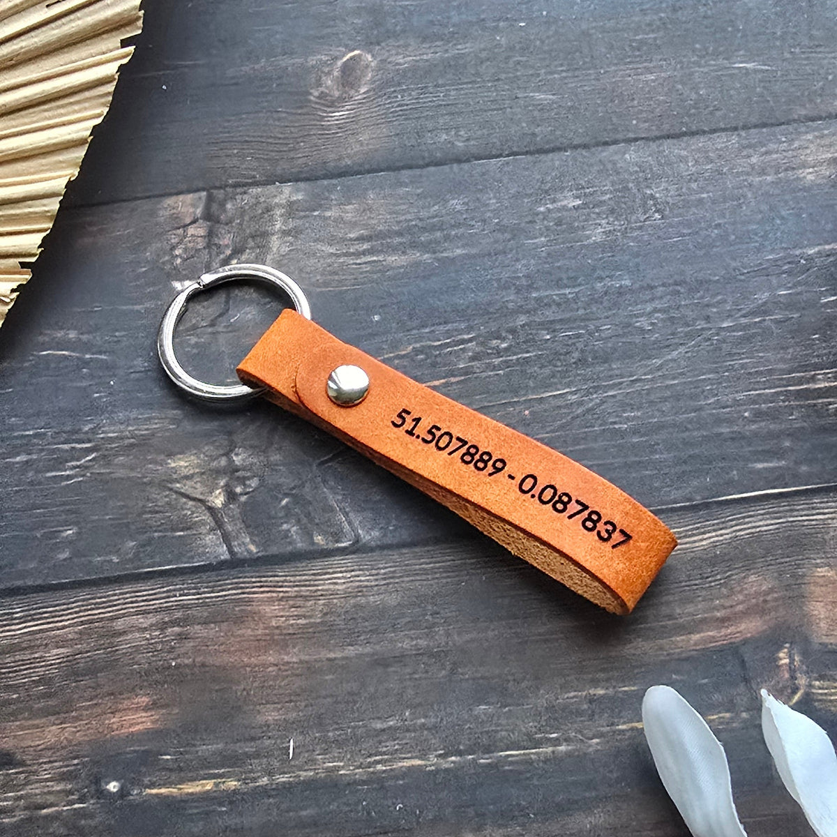 A personalized orange leather keychain with a silver split ring, featuring a printed phone number and a silver rivet, displayed on a dark wood background with decorative props.