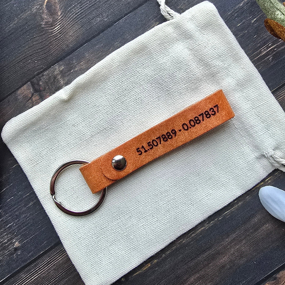 A personalized leather keychain with a metal key ring, resting on a small off-white fabric pouch over a dark wood surface. The leather tab is tan and features black printed text and a silver rivet.