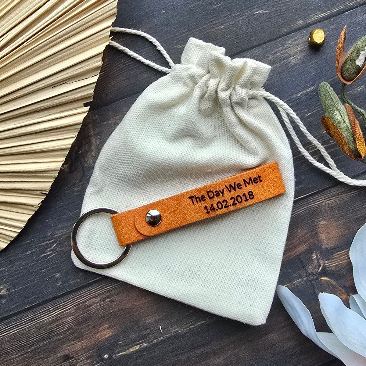 A cream-colored cotton drawstring pouch with a personalized orange leather strap keychain, styled on a dark wooden background with decorative props.
