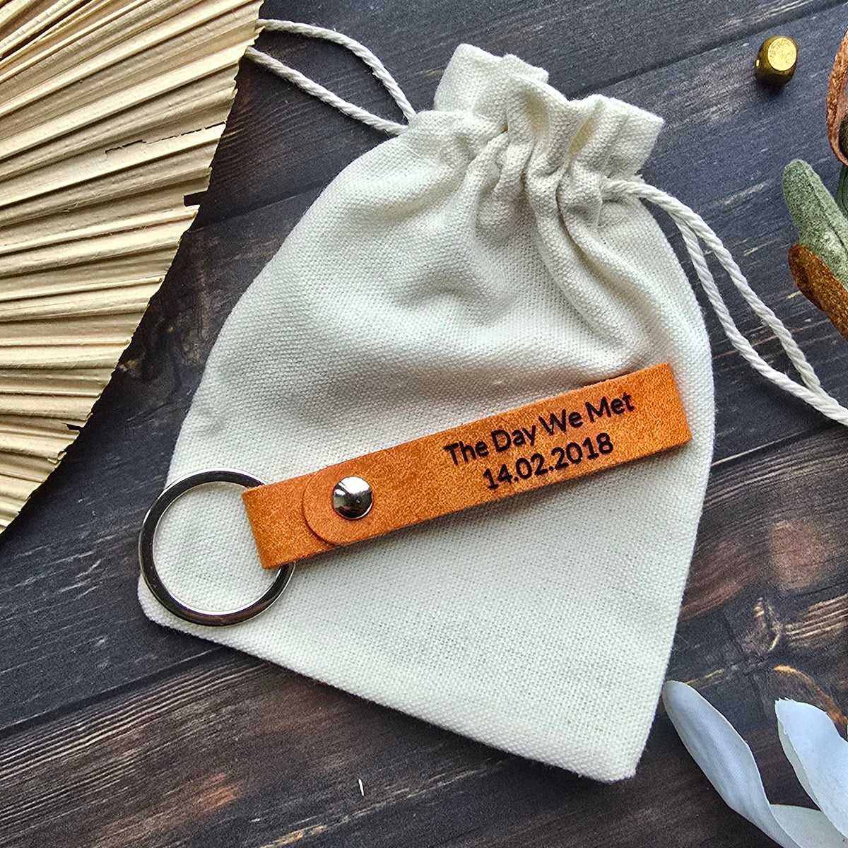 A cream-colored fabric drawstring pouch with a personalized orange leather key tag engraved with 'The Day We Meet 1.20.