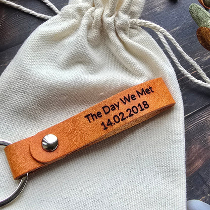 A personalized canvas drawstring gift pouch with a tan leather keychain strap engraved with 'The Day We Met 14.02.