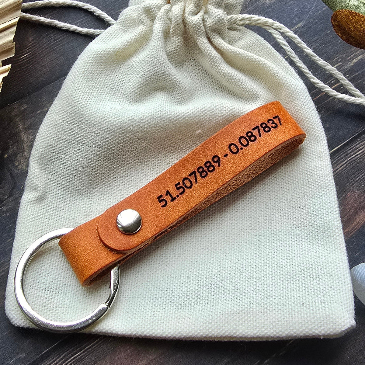 A personalized canvas drawstring pouch in cream, featuring a brown leather keychain strap with a silver key ring and a custom printed phone number, displayed on a dark wooden surface.
