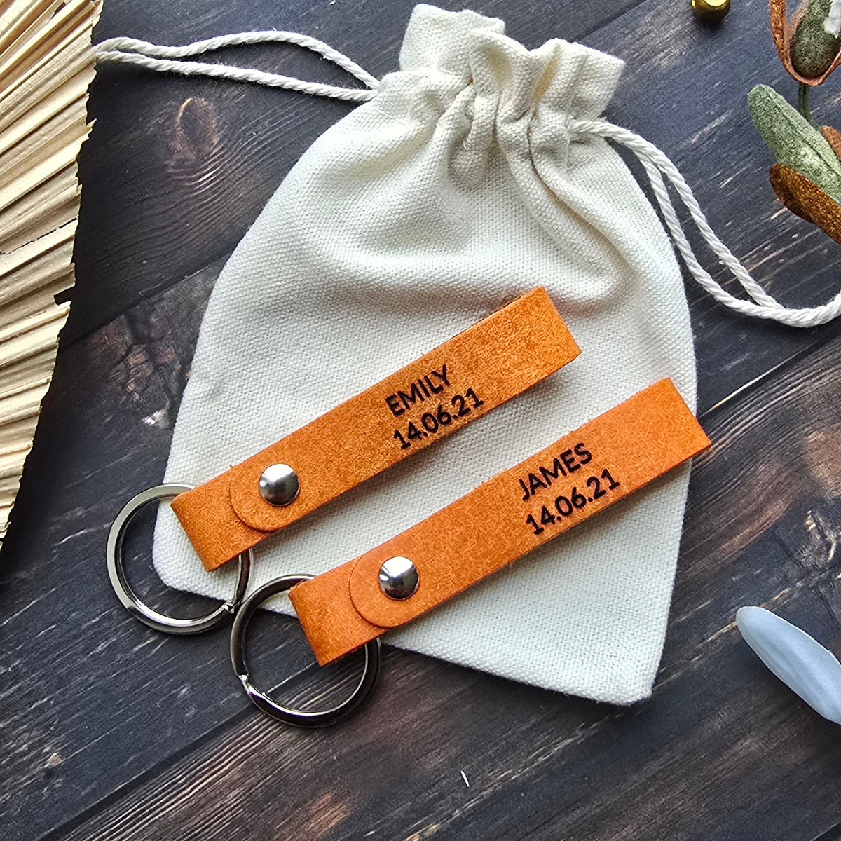 Two personalized leather keychains with orange tabs and silver key rings, displayed on a cream drawstring pouch over a dark wood background with decorative props.