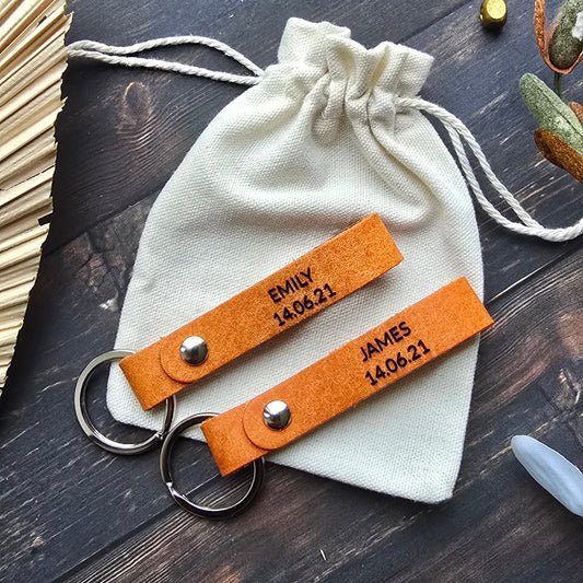 Two personalized orange leather keychains with silver key rings are displayed on a cream drawstring pouch over a dark wood background. The keychains have custom engraved names and dates.