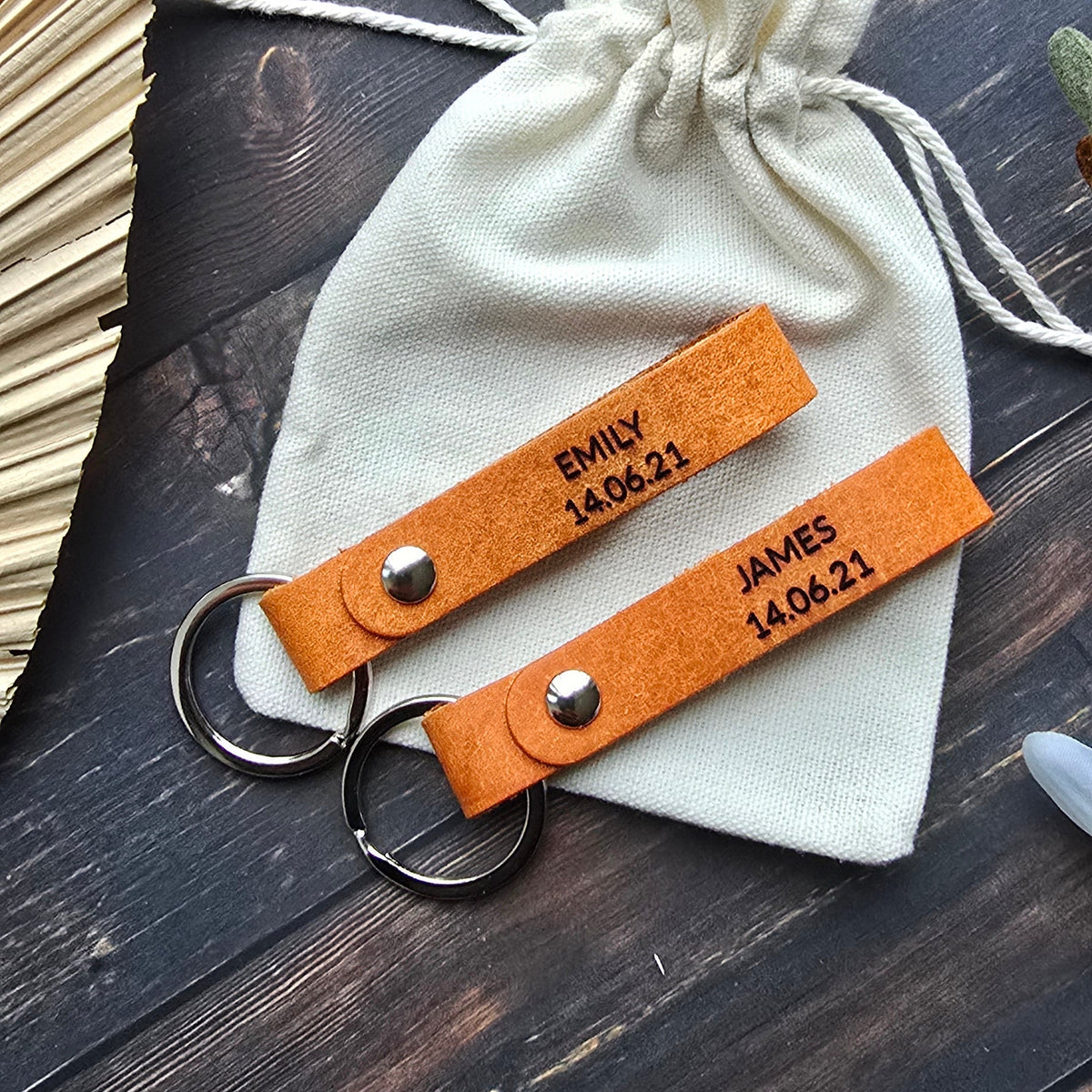 Two personalized orange leather keychains with silver key rings, displayed on a cream drawstring pouch over a dark wood background.