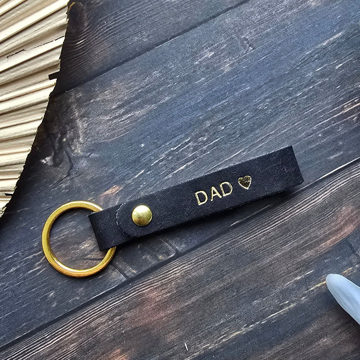 A black leather keychain with gold hardware and the word 'DAD' printed in gold, styled on a dark wooden surface with decorative props.