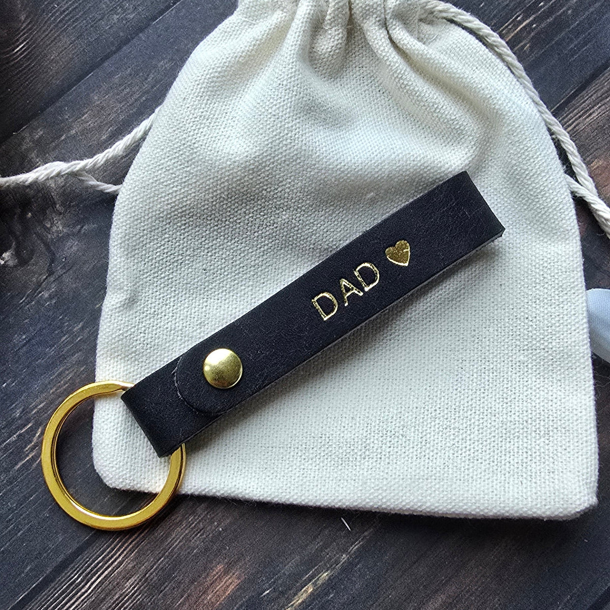 A black leather keychain with gold hardware and the word 'DAD' printed in gold, resting on a small cream drawstring pouch over a dark wooden surface.