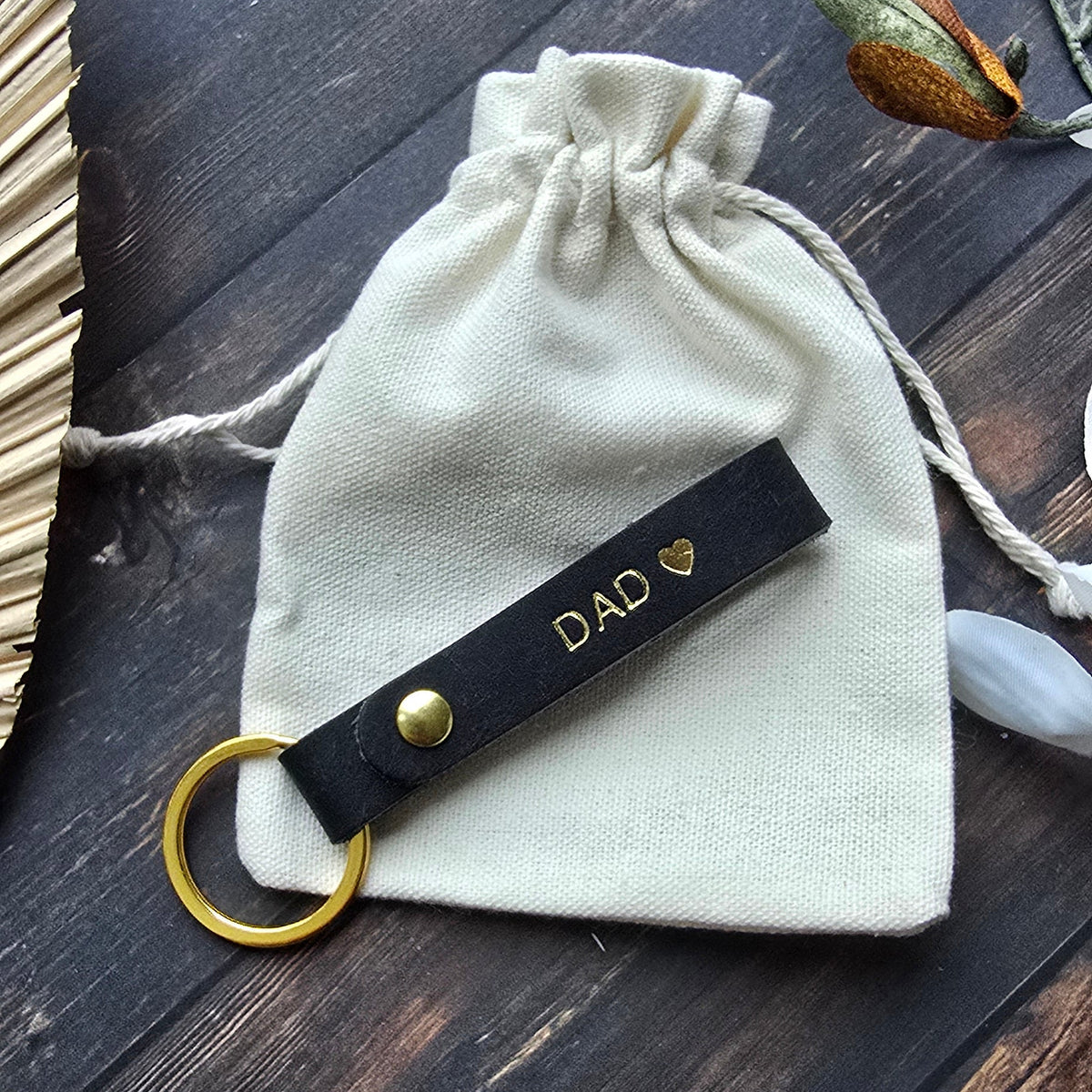 A cream-colored cotton drawstring pouch with a black leather keychain strap featuring gold hardware and the text 'DAD' in gold, displayed on a dark wood background with decorative crystals.