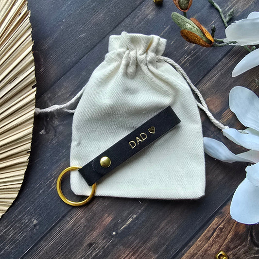 A cream-colored drawstring pouch with a black leather keychain strap featuring gold hardware and the text 'DAD ❤️', styled on a dark wooden background with decorative dried flowers and gold bells.