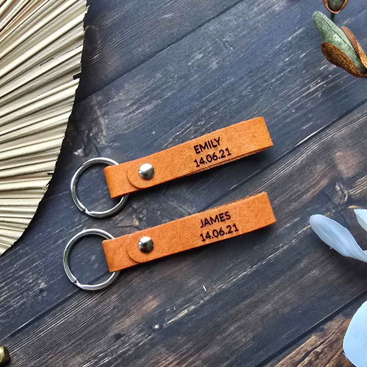 Two personalized brown leather keychains with engraved names and dates, attached to silver key rings, displayed on a dark wooden surface with decorative props.
