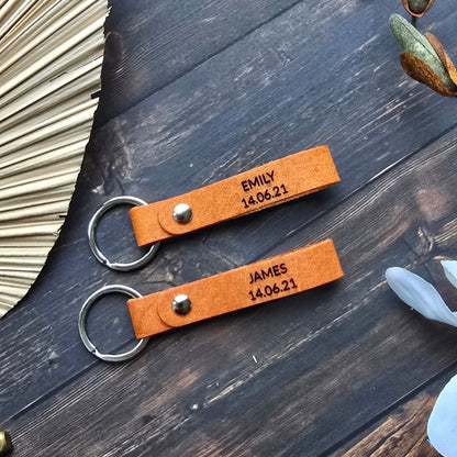 Two personalized brown leather keychains with engraved names and dates, attached to silver key rings, displayed on a dark wooden surface with decorative props.