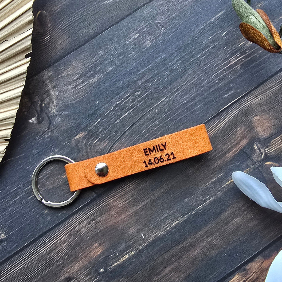 A close-up flat lay of a small orange leather keychain with a metal split ring, printed with the text 'EMAY 14.06.