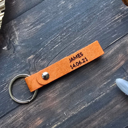 A personalized orange leather keychain with a metal split ring, featuring the engraved text 'JAMES 16:04-23', displayed on a dark rustic wooden background with decorative props.