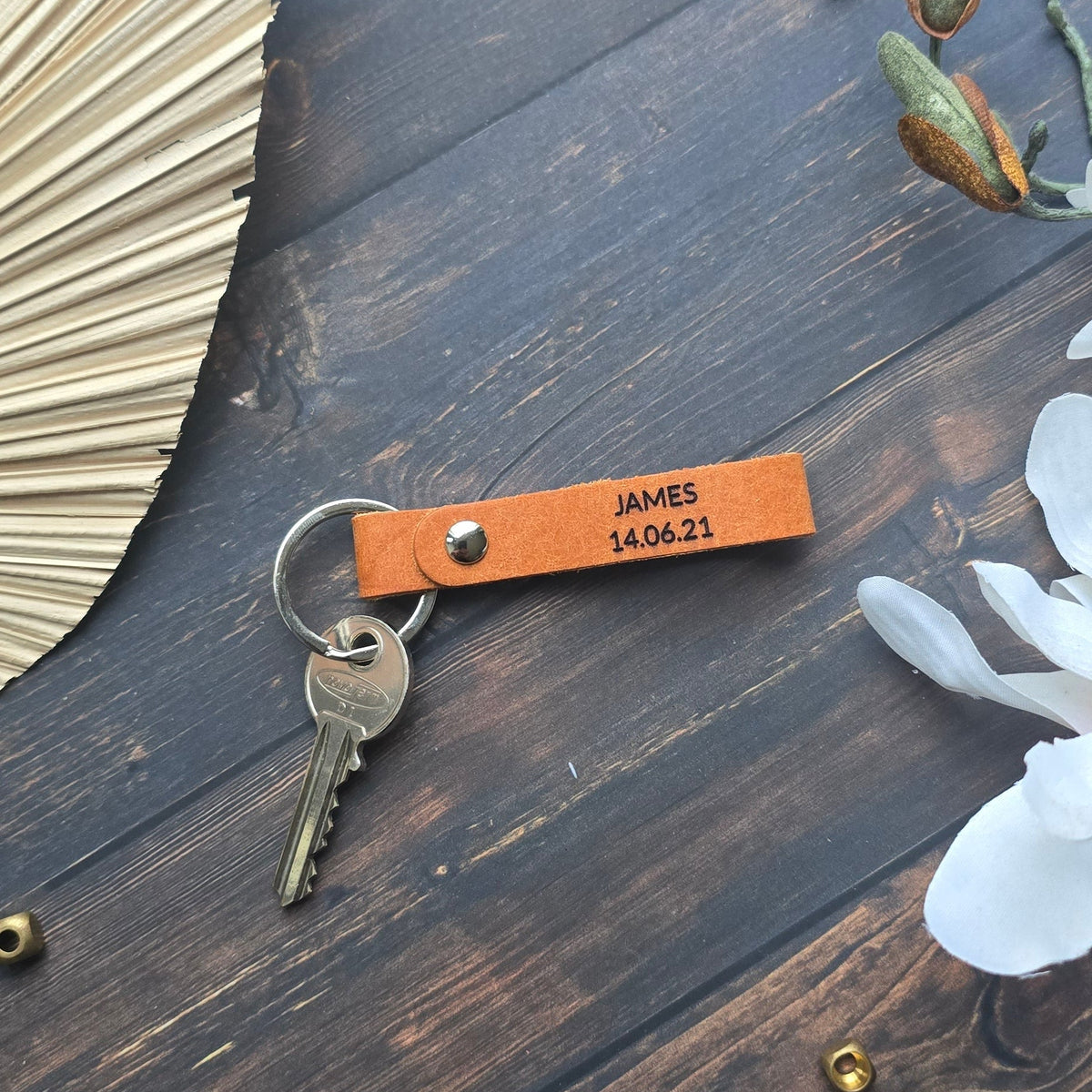 A personalized orange leather keychain with the name 'JAMES HARRIS' printed on it, attached to a metal key ring with keys, styled on a dark wood background with dried flowers and small brass pins.