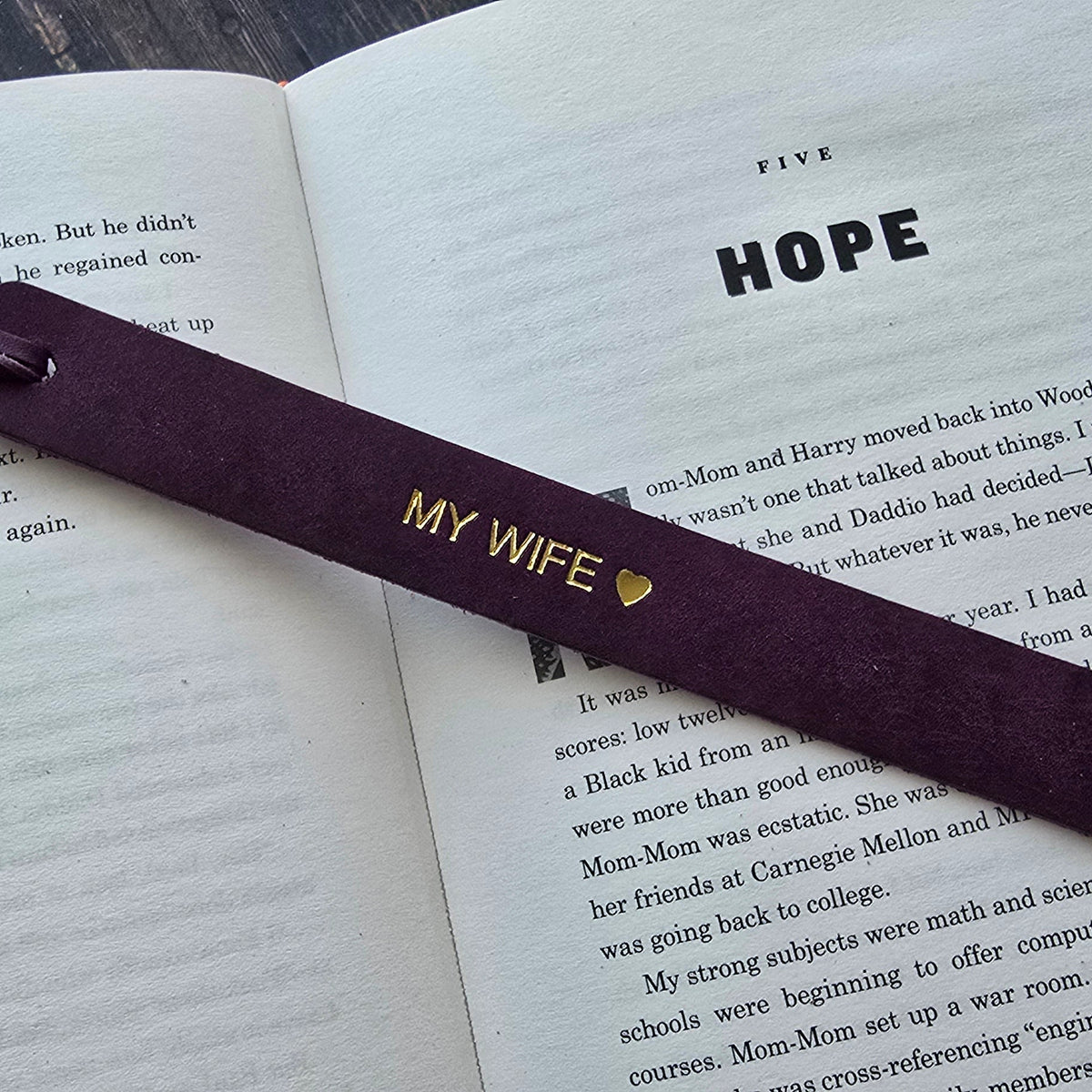 A dark purple wooden bookmark with gold lettering reading 'MY WIFE' and a small heart, resting on an open book with the word 'HOPE' visible on the page.