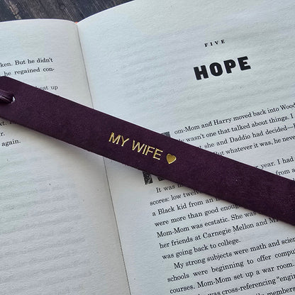 A dark purple wooden bookmark with gold lettering reading 'MY WIFE' and a small heart, resting on an open book with the word 'HOPE' visible on the page.
