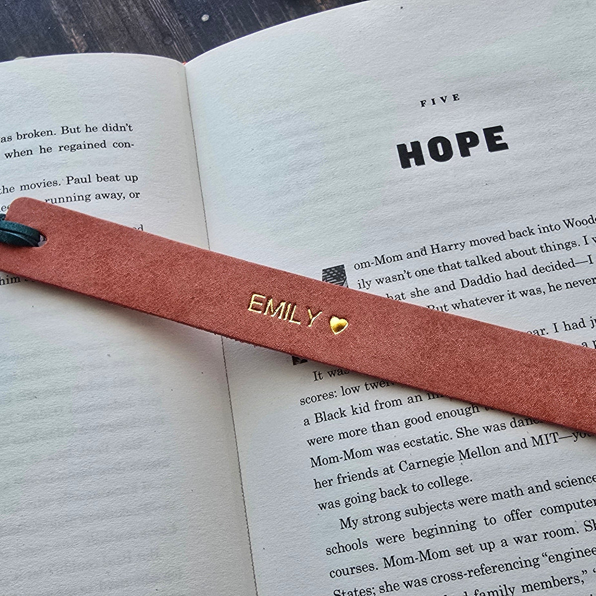 A pink leather pen with gold lettering and a blue ink tip rests on an open book, with the name 'EMILY' and a heart engraved on the front.