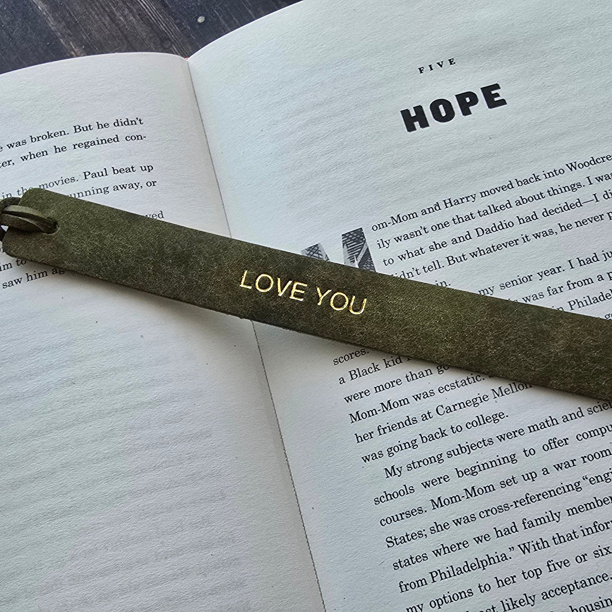 A dark olive green wooden bookmark with 'LOVE YOU' engraved in gold, resting on an open book with the word 'HOPE' on the page, set on a dark wood background.