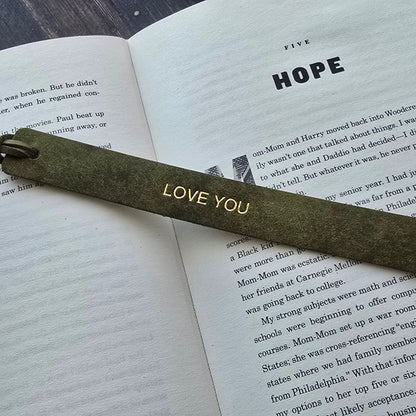 A dark olive green wooden bookmark with 'LOVE YOU' engraved in gold, resting on an open book with the word 'HOPE' on the page, set on a dark wood background.