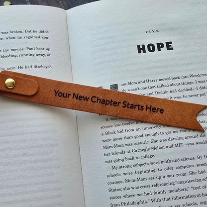 A brown leather bookmark shaped like a ribbon bookmark with a gold button, placed on an open book. The bookmark has placeholder text that reads 'Your New Chapter Starts Here'.