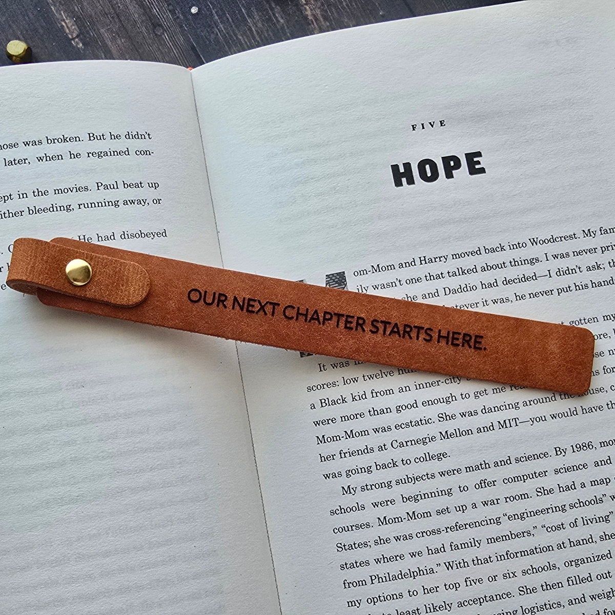 A brown leather bookmark with gold hardware rests across an open book on a dark wooden tabletop. The bookmark has the text 'OUR NEXT CHAPTER STARTS HERE' printed on it.