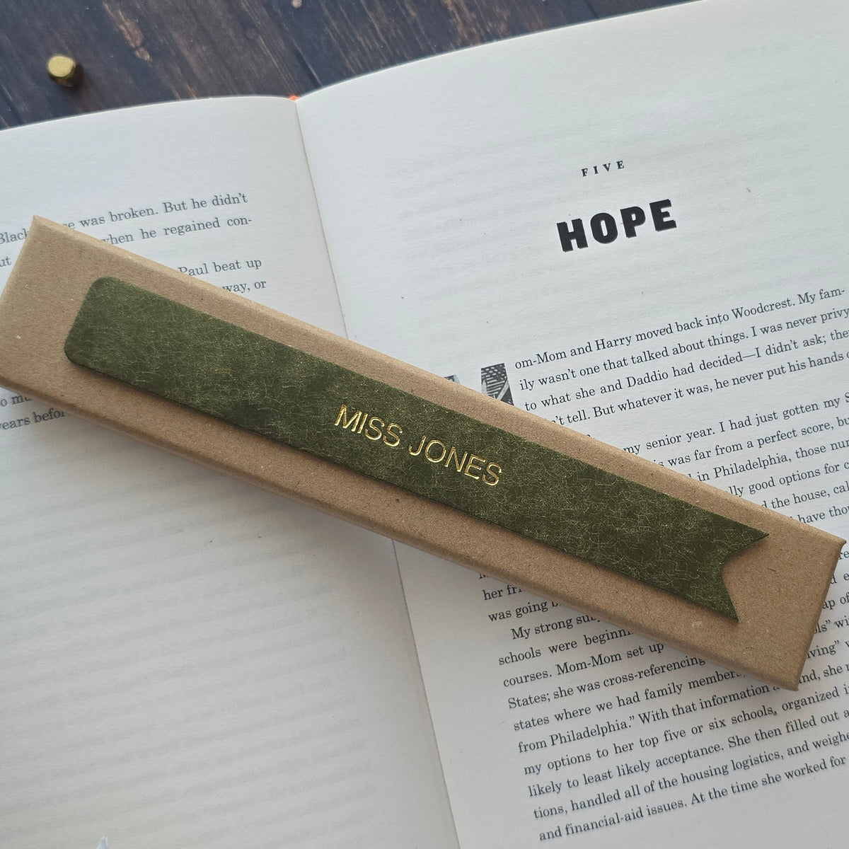 A rectangular kraft-paper bookmark with a green leather bookmark reading 'MISS JONES' in gold lettering