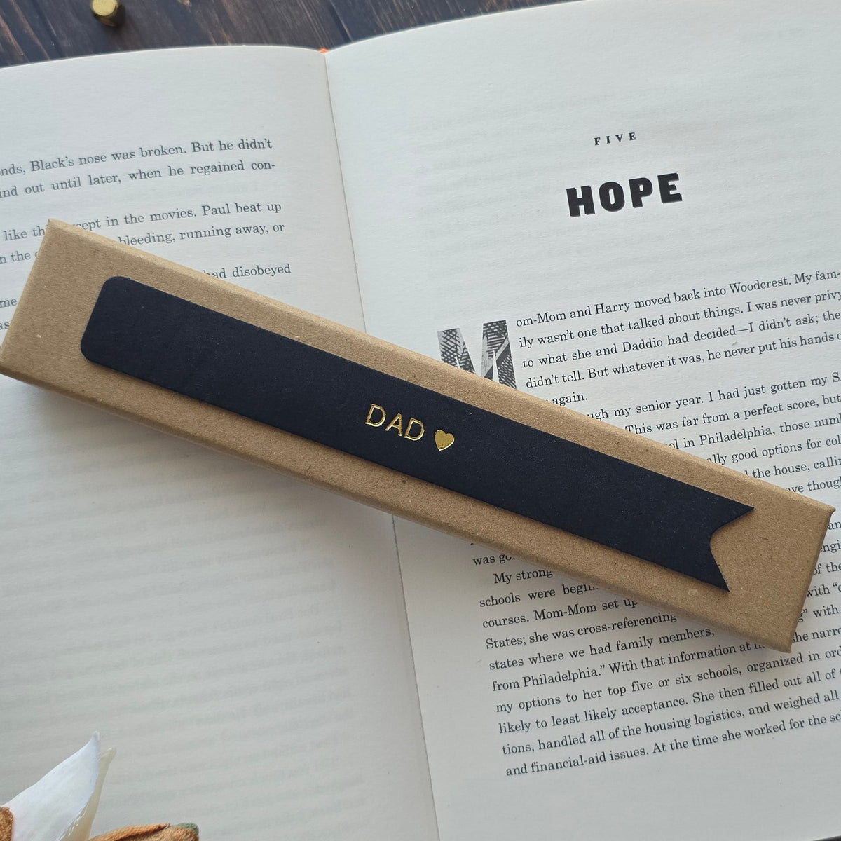 A black leather bookmark with the word 'DAD' and a small gold heart printed on it, resting on an open book. 