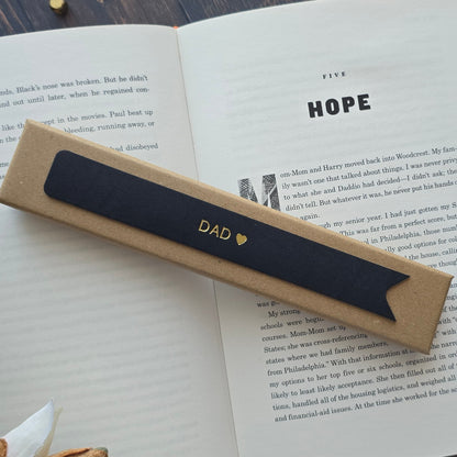 A black leather bookmark with the word 'DAD' and a small gold heart printed on it, resting on an open book. 