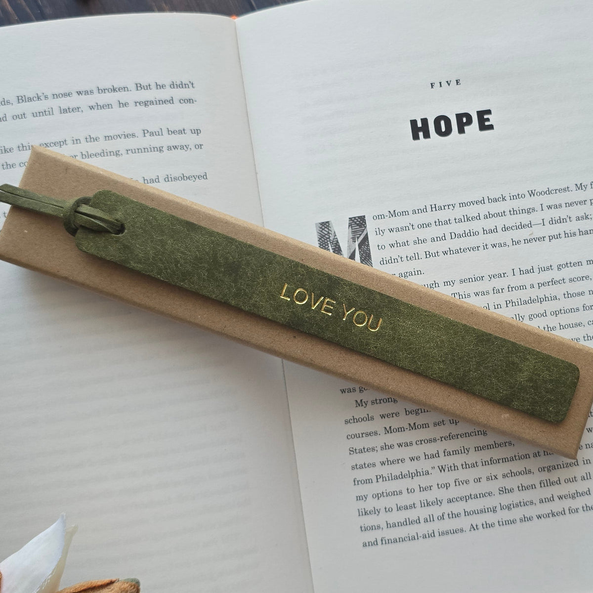 A green textured ribbon bookmark with 'LOVE YOU' printed in gold, resting on an open book. 