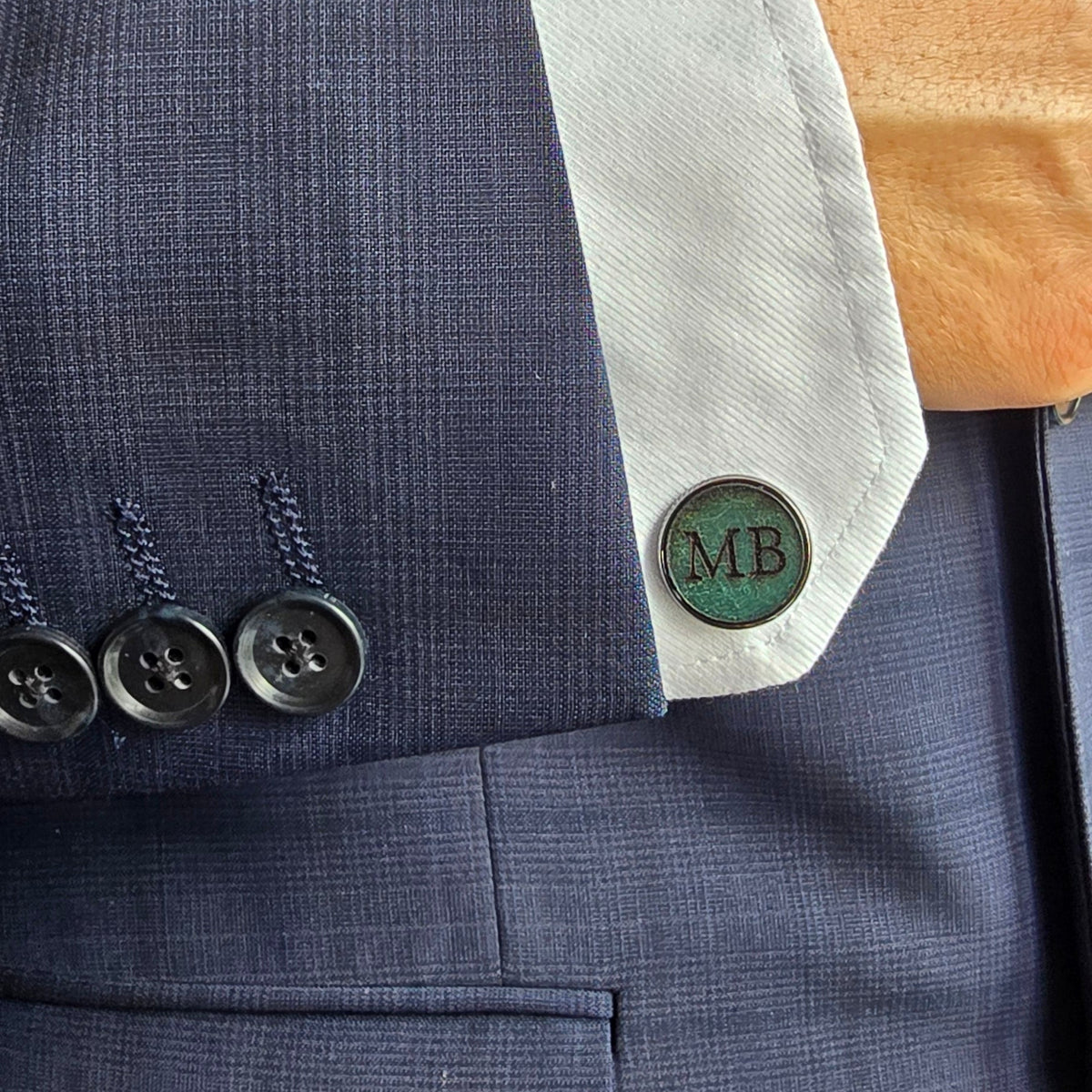 Close-up of a navy blue textured suit jacket with a white dress shirt cuff and a single round blue cufflink engraved with 'MB'.