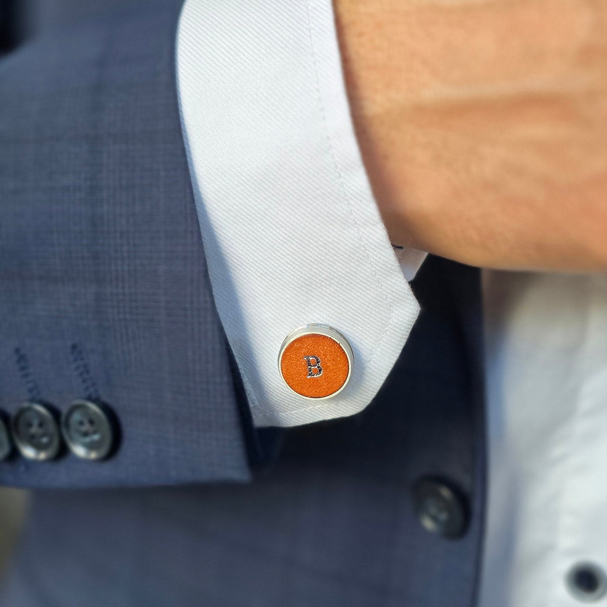 A close-up of a man's wrist and cuff as he adjusts his dress shirt cuff. The cuff features a round orange and silver cufflinks  with initials
